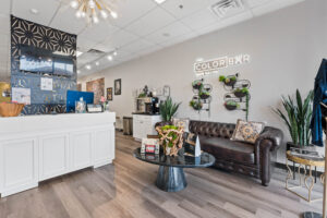 - The Color Bar, TX Modern hair salon interior with a white reception desk, brown leather sofa, decorative plants, wall art, and a neon sign reading "The Color Bar Hair Salon," where expert hair stylists craft beautiful hair highlights. - The Color Bar, TX