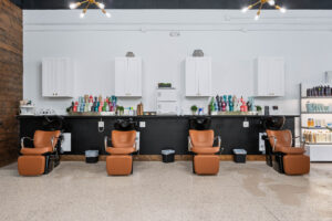 - The Color Bar, TX Three brown salon chairs with black wash stations in front of a counter holding hair products, perfect for your next haircut or balayage, set against a wall with white cabinets and shelves. - The Color Bar, TX
