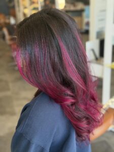 - The Color Bar, TX Person with long, dark brown hair featuring vibrant magenta balayage highlights, styled in loose waves, wearing a dark-colored top in a hair salon setting. - The Color Bar, TX