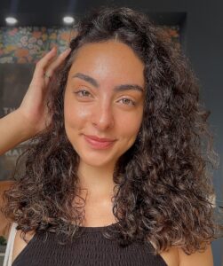 - The Color Bar, TX A woman with curly brown hair and a black top smiles at the camera, standing in front of a dark background with a decorative floral pattern, her balayage highlights adding dimension to her vibrant hairstyle. - The Color Bar, TX