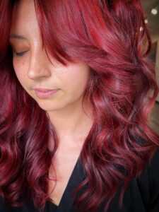 - The Color Bar, TX A person with long, wavy, vibrant red hair featuring subtle highlights and side-swept bangs looks downward with eyes closed, wearing a black top. - The Color Bar, TX