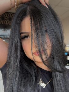 - The Color Bar, TX A woman with long, straight black hair and subtle balayage partially covering her face looks at the camera, wearing a black top, gold jewelry, and a nameplate necklace. - The Color Bar, TX