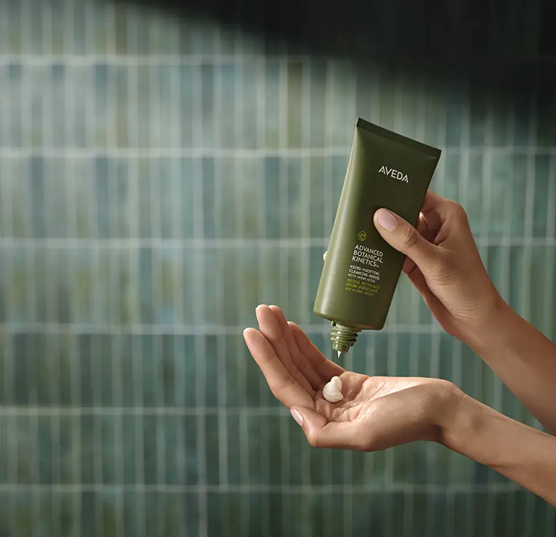 - The Color Bar, TX A person dispenses a cream from a green Aveda tube onto their hand in front of a green tiled background. - The Color Bar, TX