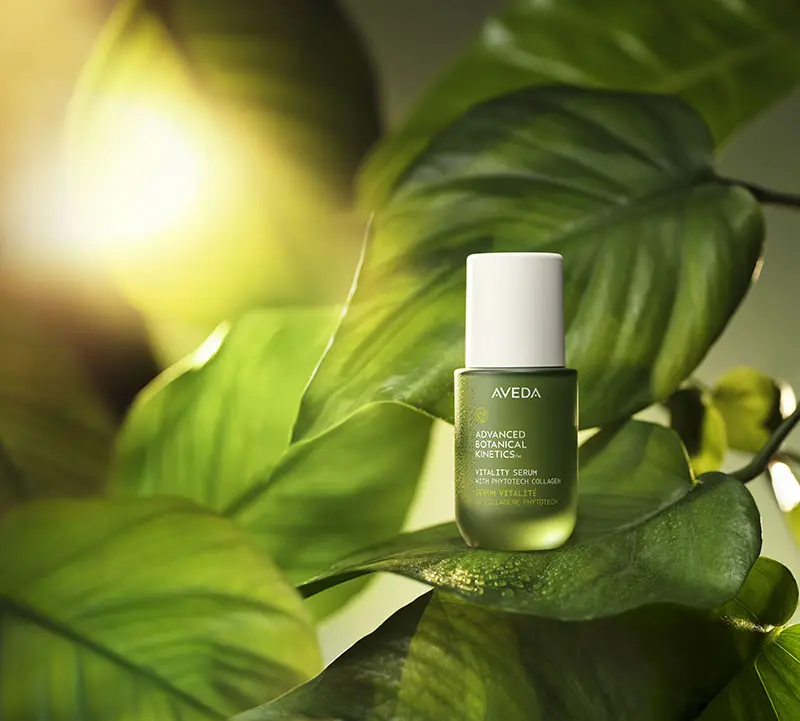 - The Color Bar, TX A green bottle of Aveda Advanced Botanical Kinetics serum is displayed on large green leaves with sunlight in the background. - The Color Bar, TX
