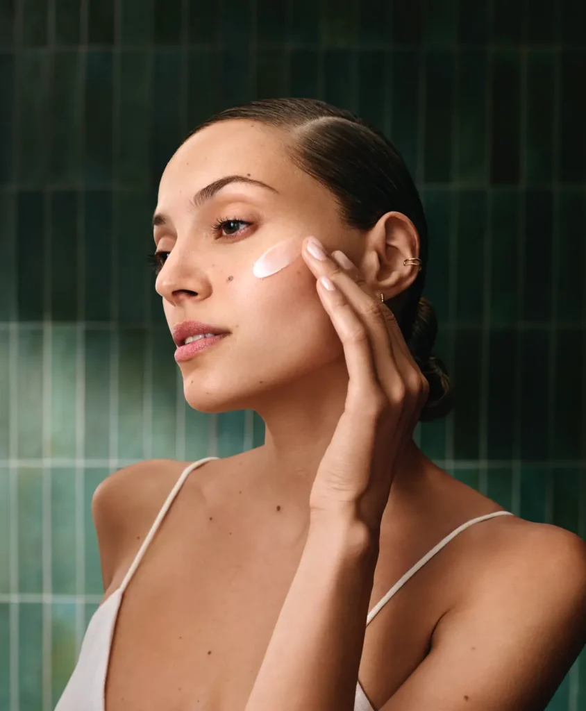 - The Color Bar, TX A woman in a white camisole applies Aveda Botanical Kinetics skincare cream to her cheek with her fingers, set against a green tiled background. - The Color Bar, TX