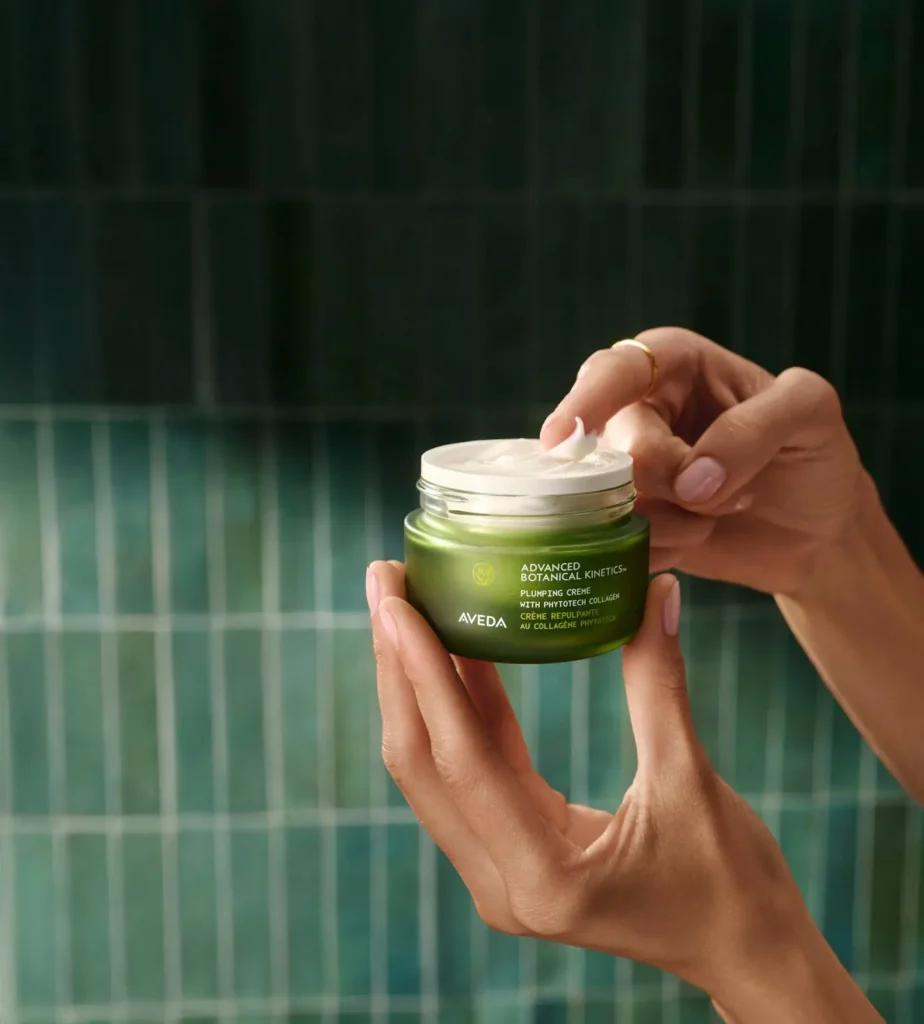 - The Color Bar, TX A person holds an open jar of Aveda Advanced Botanical Repair Intensive Strengthening Masque, showcasing the brand's commitment to botanical skincare, in front of a green tiled background. - The Color Bar, TX