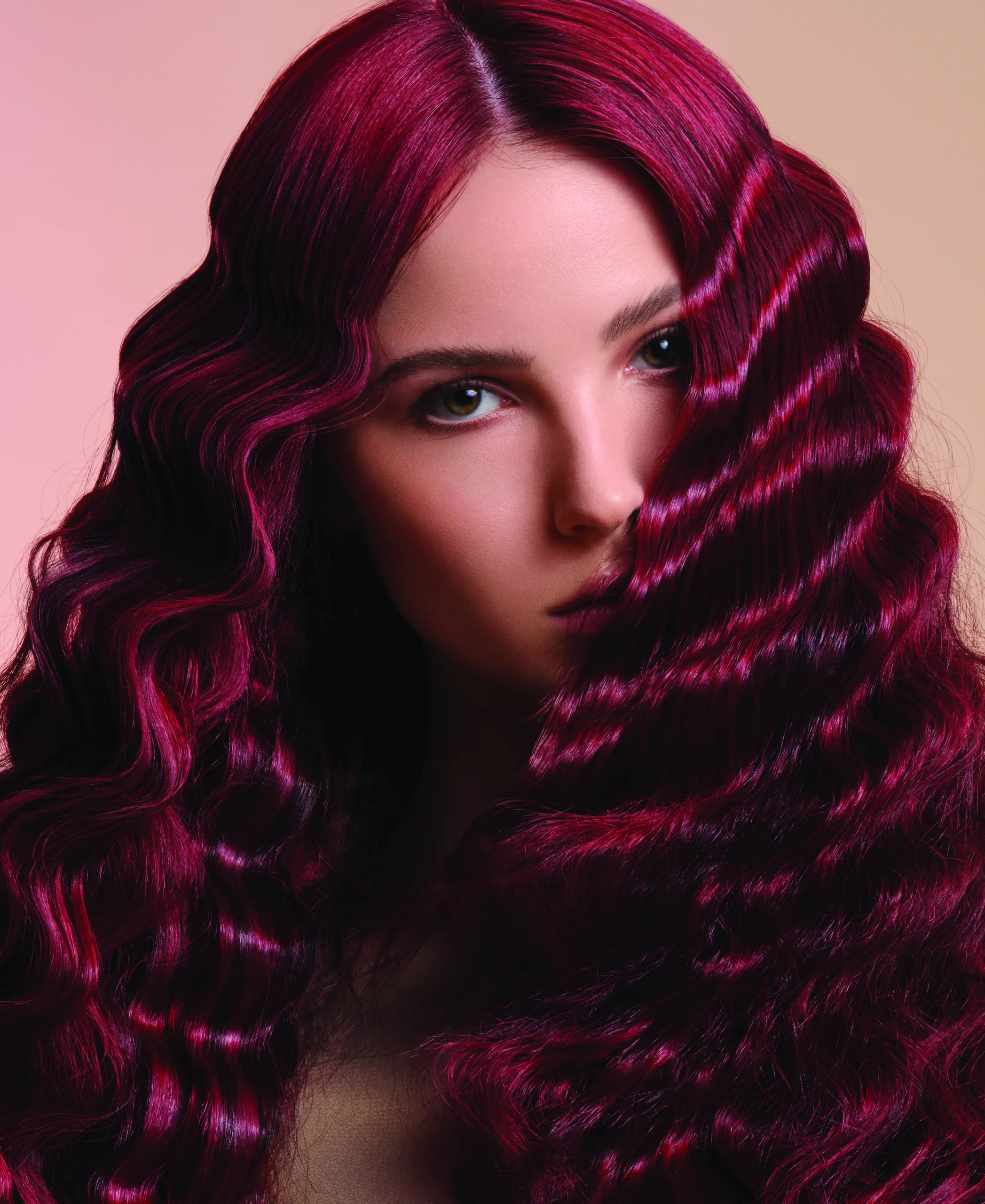 - The Color Bar, TX Woman with long, dark magenta crimped hair looks at the camera against a blurred beige and pink background. - The Color Bar, TX