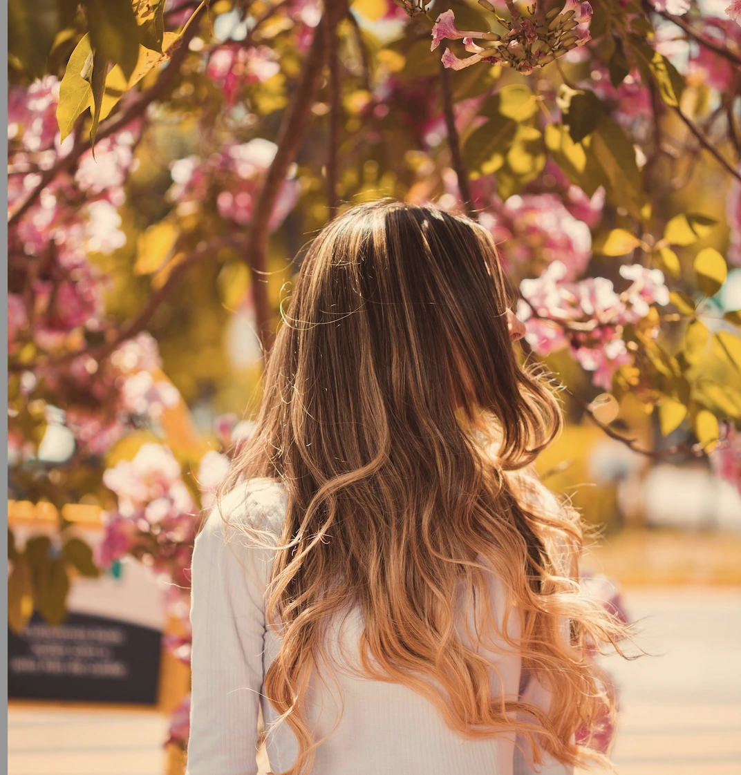 - The Color Bar, TX A person with long, wavy, blonde-brown hair stands outdoors facing away, surrounded by blooming pink flowers and green leaves. - The Color Bar, TX