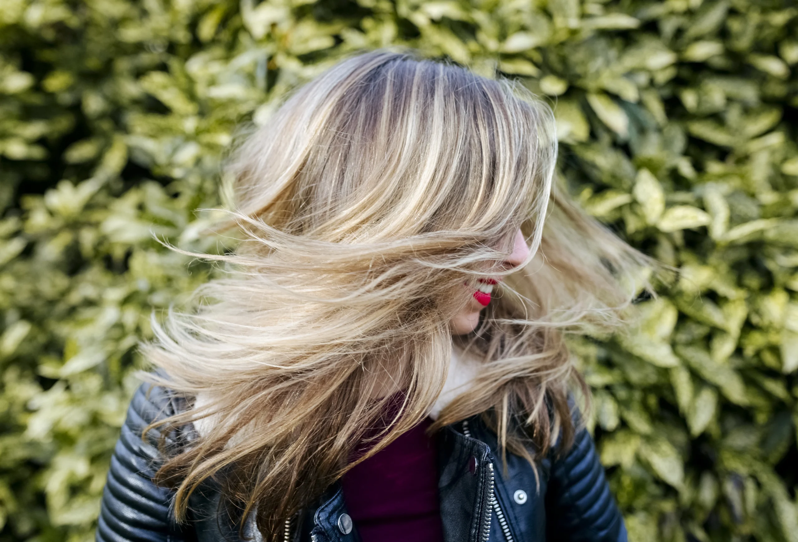 - The Color Bar, TX A person with blonde hair and a leather jacket turns their head, hair in motion, with green foliage in the background. - The Color Bar, TX