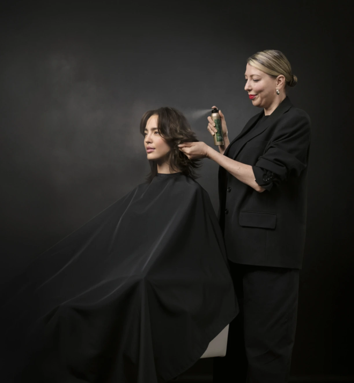 - The Color Bar, TX A hairstylist dressed in black sprays hair product on a seated client's hair, both against a plain dark background. - The Color Bar, TX