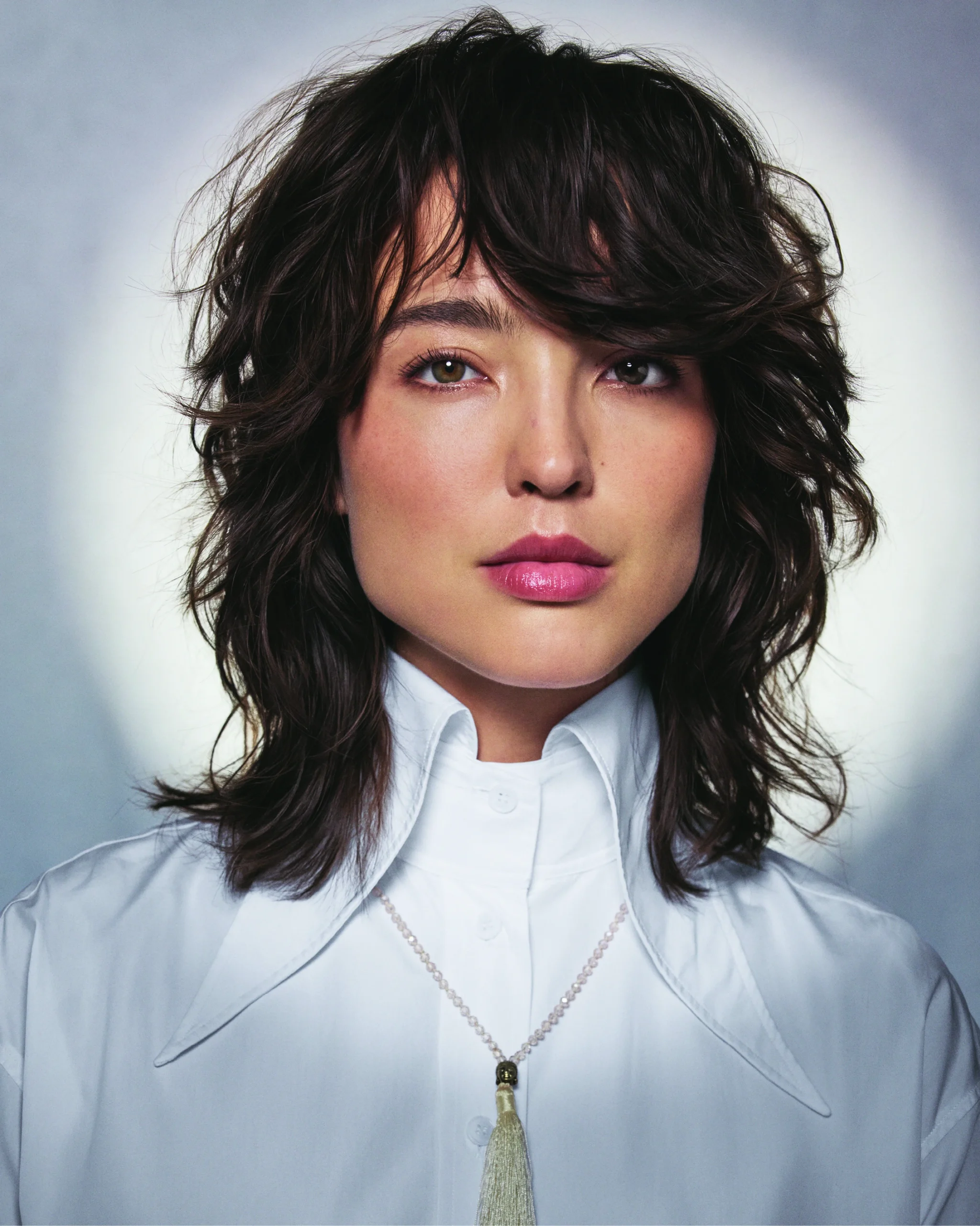 - The Color Bar, TX A person with medium-length, wavy brown hair and bangs wears a white collared shirt and a necklace with a tassel pendant, posing against a light background. - The Color Bar, TX