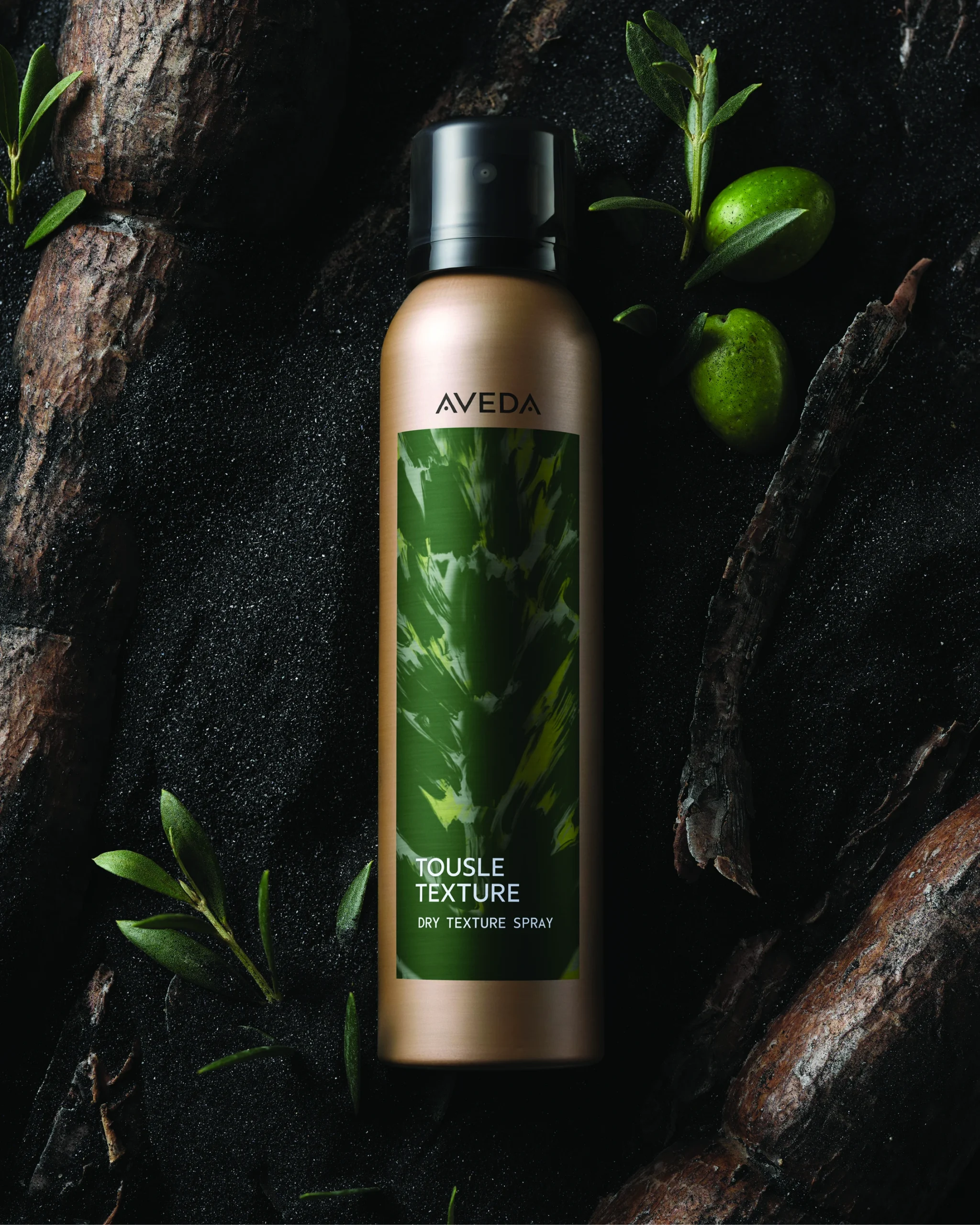 - The Color Bar, TX Aveda Tousle Texture Dry Texture Spray bottle on a dark surface with green olives and leaves scattered around. - The Color Bar, TX