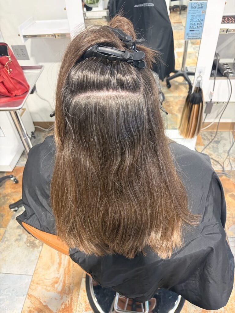 - The Color Bar, TX Person with medium-length brown hair sectioned and clipped up, sitting in a salon chair with hair tools and a mirror visible in the background. - The Color Bar, TX