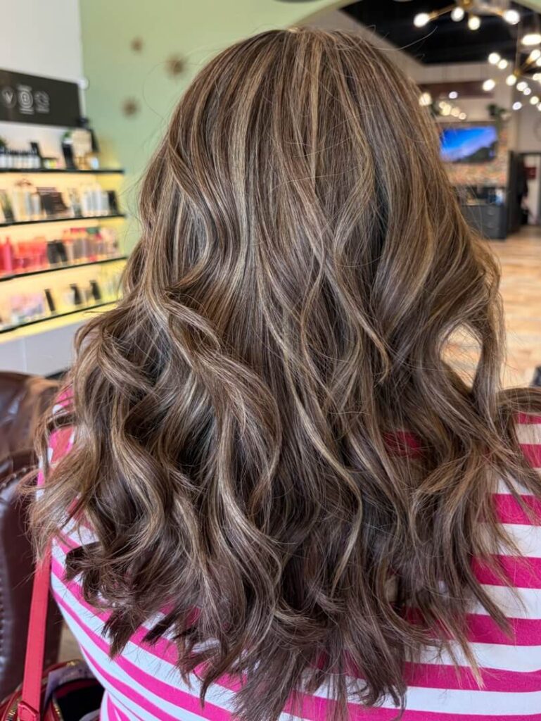 - The Color Bar, TX Person with long, wavy, light brown hair with highlights, wearing a pink and white striped top, sitting in a salon with shelves of hair products visible in the background. - The Color Bar, TX