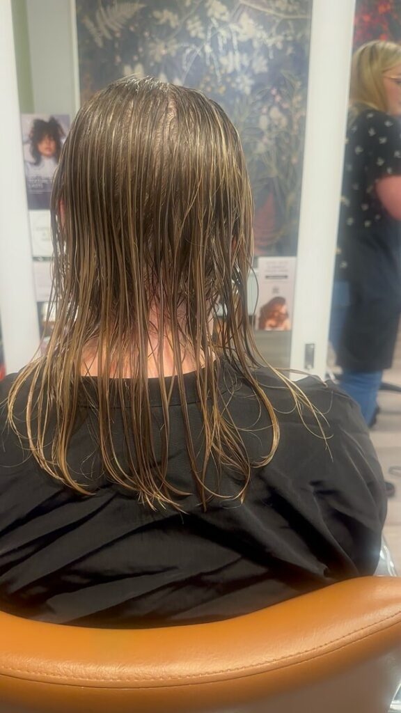 - The Color Bar, TX Person with wet, shoulder-length blond hair sits in a salon chair facing a mirror; a hairdresser stands in the background. - The Color Bar, TX